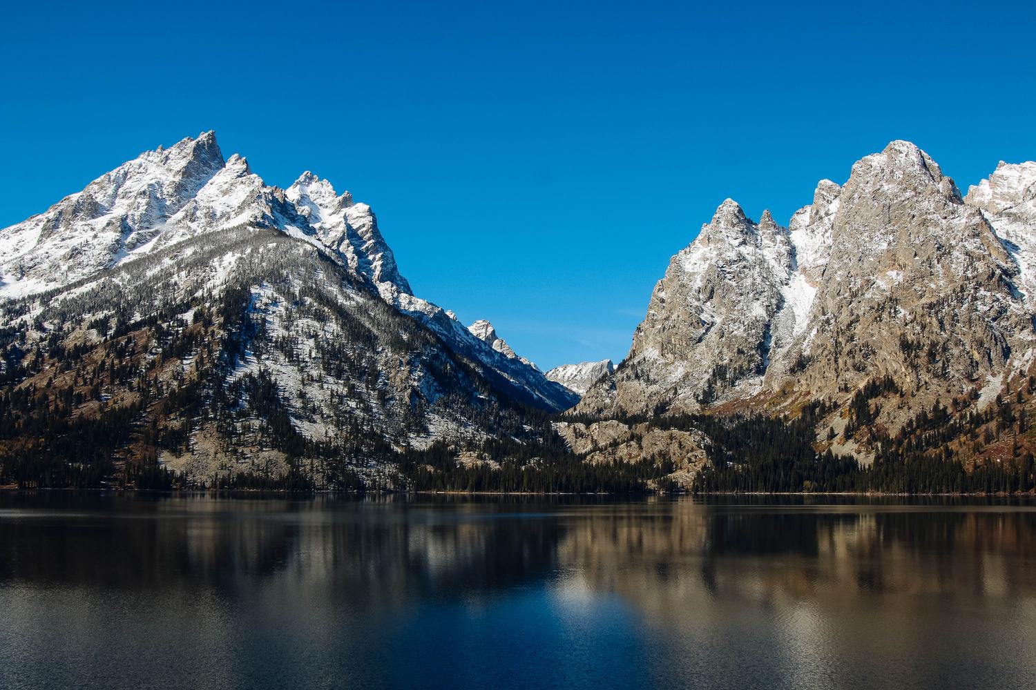 Snow streaked Teton peaks rising straight out of the dark blue water of Jenny Lake.