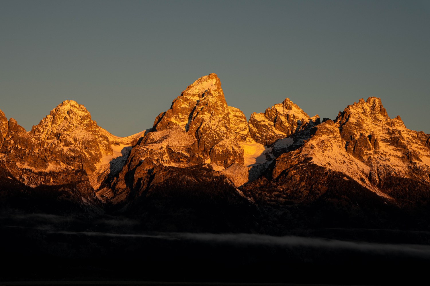 Sunrise lighting the jagged peaks of the Teton Range orange above dark forested slopes.